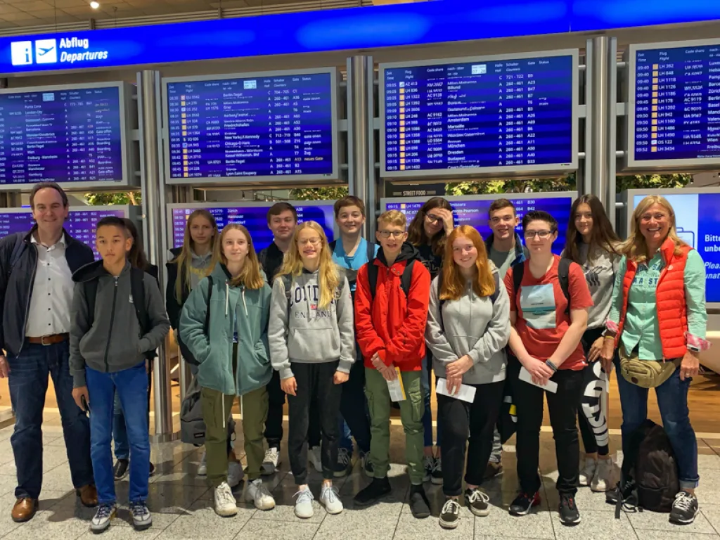 Die Reisegruppe steht vor der Anzeigetafel am Flughafen.